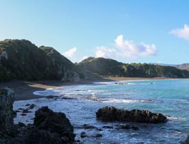 Breaker Bay on a sunny day, blue and green waves crashing on the stoney shore, with green cliffs surrounding.