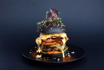 A charcoal milk bun with sesame seeds contains a chorizo and beef patty, bacon, hash, fried egg and oozing cheese. Microgreens sit atop with a sprinkling of paprika on a black plate.