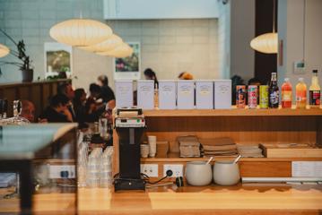 Behind the counter at Twenty Eight, a café in Lower Hutt. There are tea boxes, other beverages on the top shelf, napkins and takeaway containers stacked in the middle, water glasses, a coffee machine and two bowls on the wood surface.