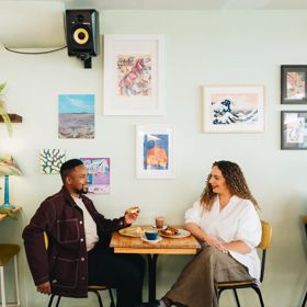 Two people sit at a table inside Swimsuit on Dixon Street, talking and drinking coffee. The wall behind them is duck egg green, and the wall is covered in art.
