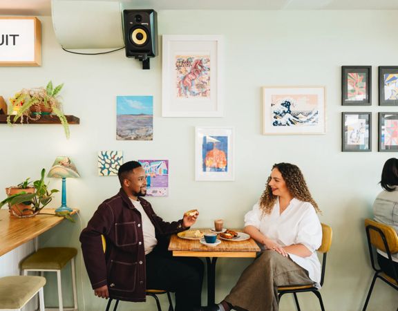 Two people sit at a table inside Swimsuit on Dixon Street, talking and drinking coffee. The wall behind them is duck egg green, and the wall is covered in art.