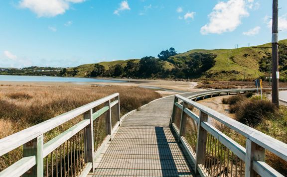 Wooden boardwalk curving through tussocks next to Pāuatahanui inlet. Blue sky.