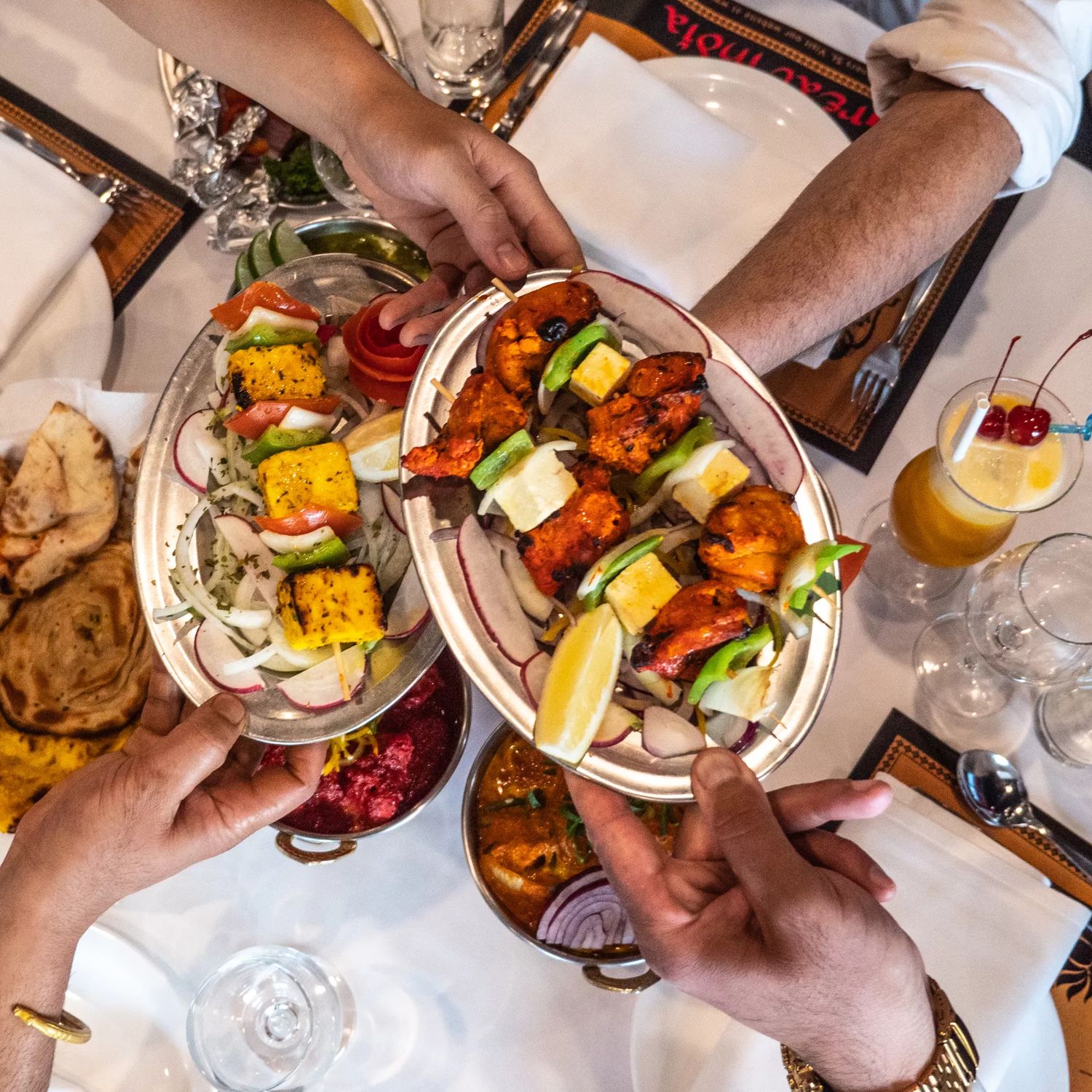 Overhead view of a shared Indian meal at Great India in Wellington, with diners’ hands reaching in to serve skewers of grilled meat and vegetables, naan bread, and assorted dishes laid out on a table.