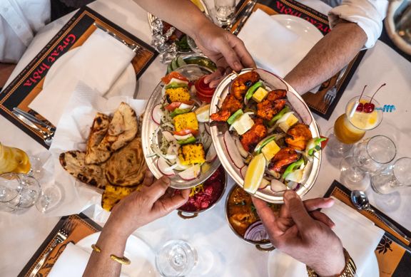 Overhead view of a shared Indian meal at Great India in Wellington, with diners’ hands reaching in to serve skewers of grilled meat and vegetables, naan bread, and assorted dishes laid out on a table.
