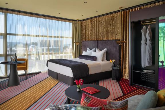 Located in Wellington's Naumi Hotel, the room features a king-sized bed, a wardrobe, and a small table with a chair. The floor-to-ceiling window, draped with sheer material, offers a city view.