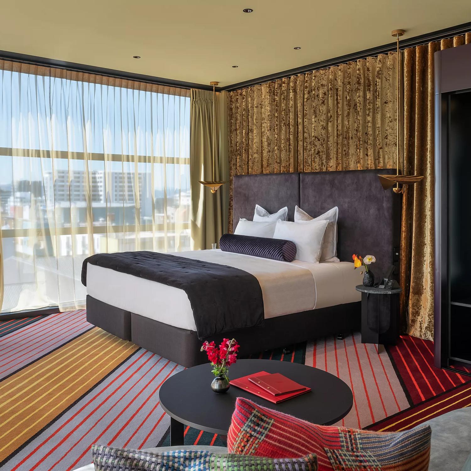 Located in Wellington's Naumi Hotel, the room features a king-sized bed, a wardrobe, and a small table with a chair. The floor-to-ceiling window, draped with sheer material, offers a city view. 