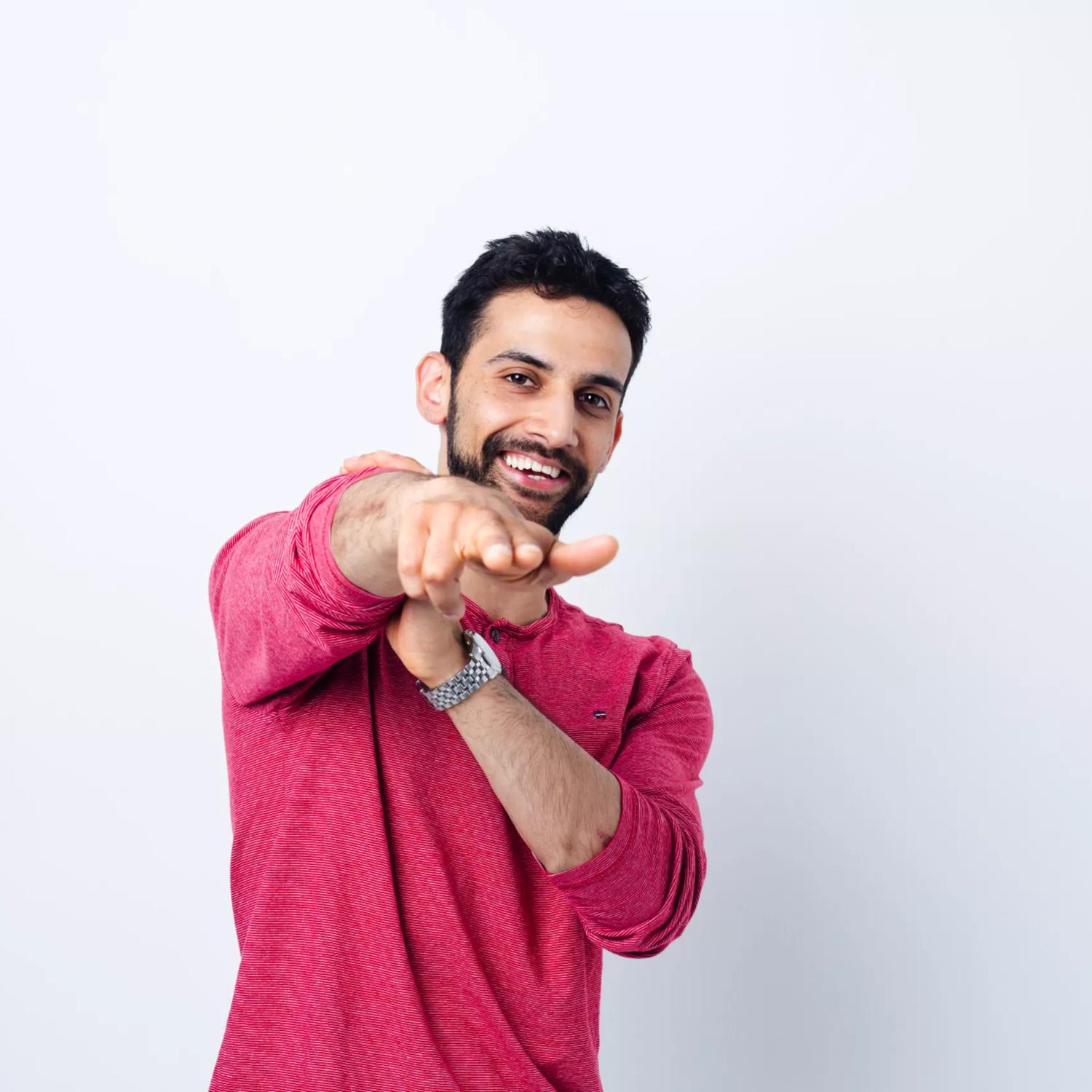 Stand-up comedian, Advait Kirtikar, wears a dark pink long sleeve shirt and poses with his right hand pointing forward. 