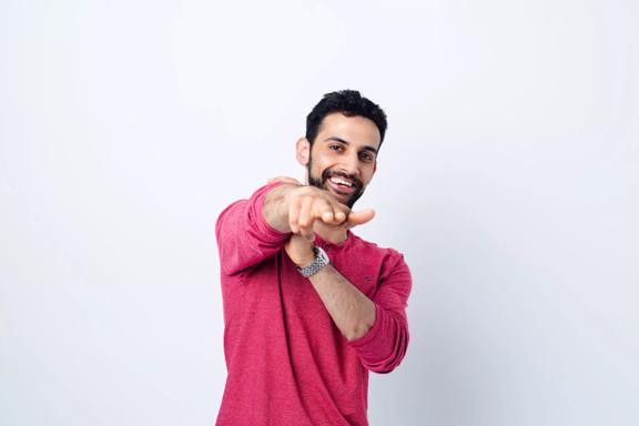 Stand-up comedian, Advait Kirtikar, wears a dark pink long sleeve shirt and poses with his right hand pointing forward. 