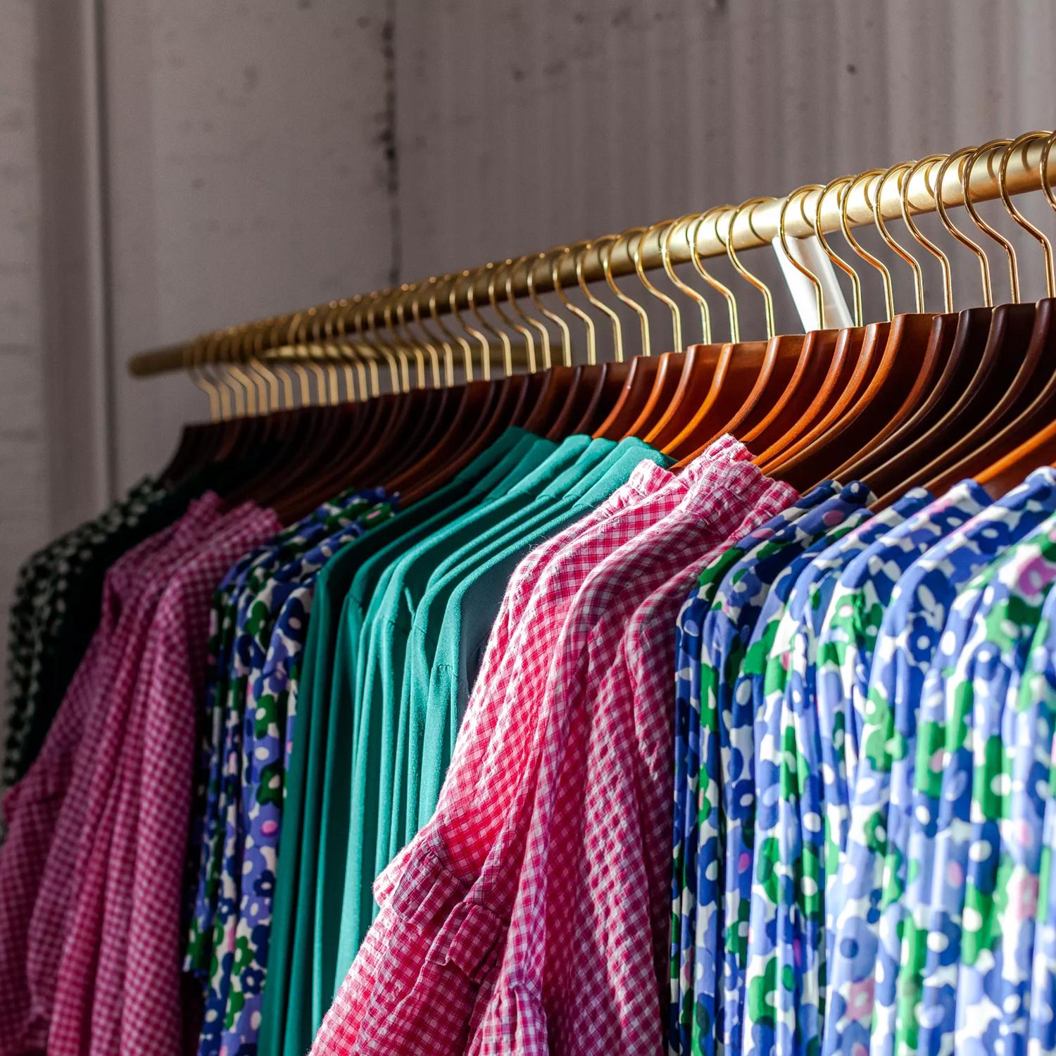 Colour coordinated clothing on wood and gold hangers lined perfectly on a rack in Twenty Seven Names.