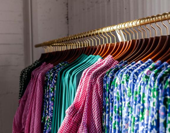 Colour coordinated clothing on wood and gold hangers lined perfectly on a rack in Twenty Seven Names.