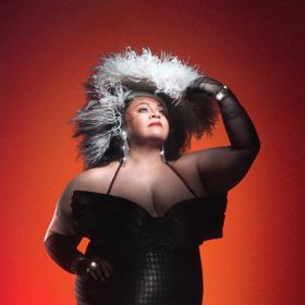 Promotional portrait for Sunset Boulevard featuring a performer in a black corseted gown, long black gloves, and a dramatic feathered headpiece, posed against a red-to-black gradient background.
