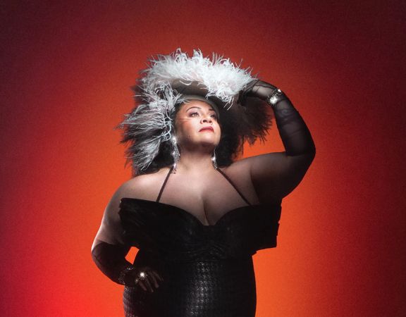 Promotional portrait for Sunset Boulevard featuring a performer in a black corseted gown, long black gloves, and a dramatic feathered headpiece, posed against a red-to-black gradient background.