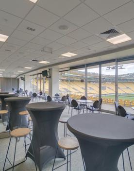 A bar set up inside the Hnry Stadium Function Centre, each bar has 4 chairs and there is around 20 tables. A large glass window looks over the field and stands.