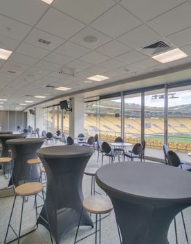 A bar set up inside the Hnry Stadium Function Centre, each bar has 4 chairs and there is around 20 tables. A large glass window looks over the field and stands.