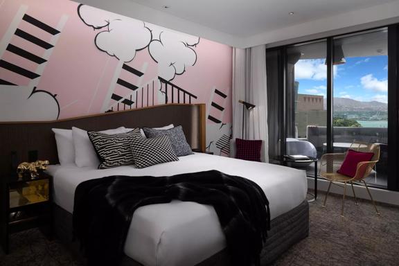 The interior of a room at the QT Wellington Hotel with a queen-sized bed, a pink wall with black and white illustrations of clouds and ladders, glass sliding doors leading to a balcony and a view of Te Papa museum, the harbour and hills in the distance.