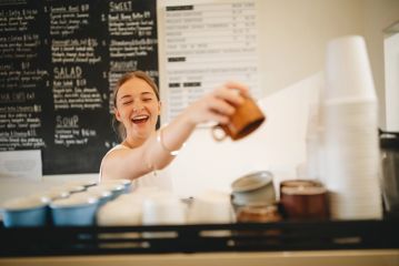 A barista grabs a mug from the coffee machine and laughs.