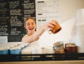 A barista grabs a mug from the coffee machine and laughs.