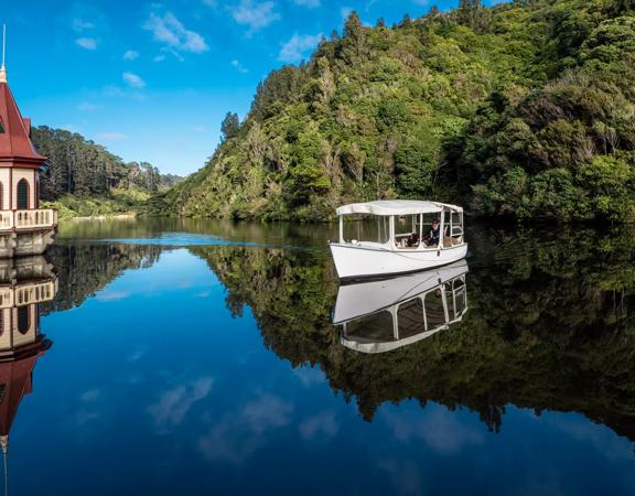 A white boat sails on the Lower Lake at Zealandia, surrounded by green bush and overhanging fern trees.