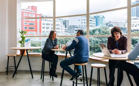 Small high tables in front of big windows at BizDojo, Wellington, with people talking over work projects.