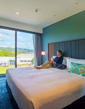 Person laying on a king bed inside The Sebel, Lower Hutt. The carpet and walls are teal and floor to ceiling windows sit on the far side of the window looking out into the Hutt.