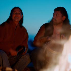 2 people sitting on Princess Bay beach at sunset, playing drums around a fire.