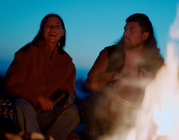 2 people sitting on Princess Bay beach at sunset, playing drums around a fire.