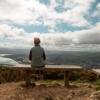 A person wearing a vest sits on a bench looking at the scenic landscape from Rangituhi/Colonial Knob Walkway.