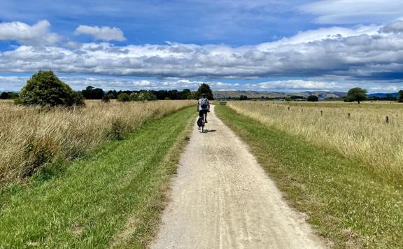 A cyclist rides on a dirt path in a field under a blue cloudy sky.