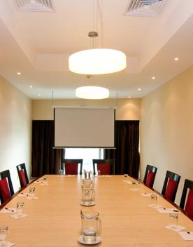Inside a Copthorne Boardroom, a large wooden table with 10 seats sits in the middle of the room, with hanging lights and a projector screen.