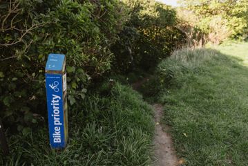 Narrow dirt pathway disappearing into the bush next to a blue 'Bike Priority' trail marker for Solitary.