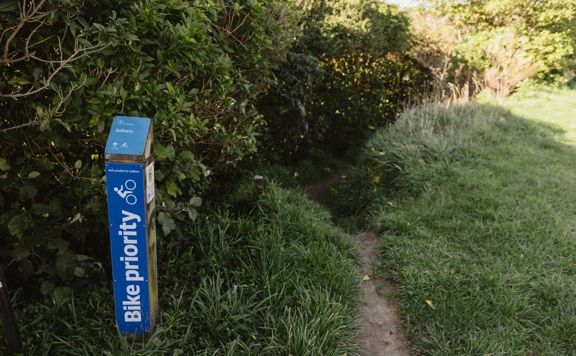 Narrow dirt pathway disappearing into the bush next to a blue 'Bike Priority' trail marker for Solitary.