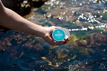 Someone holding a tin of Seasick Sunscreen Co with the surface of the water sparkling in the background.