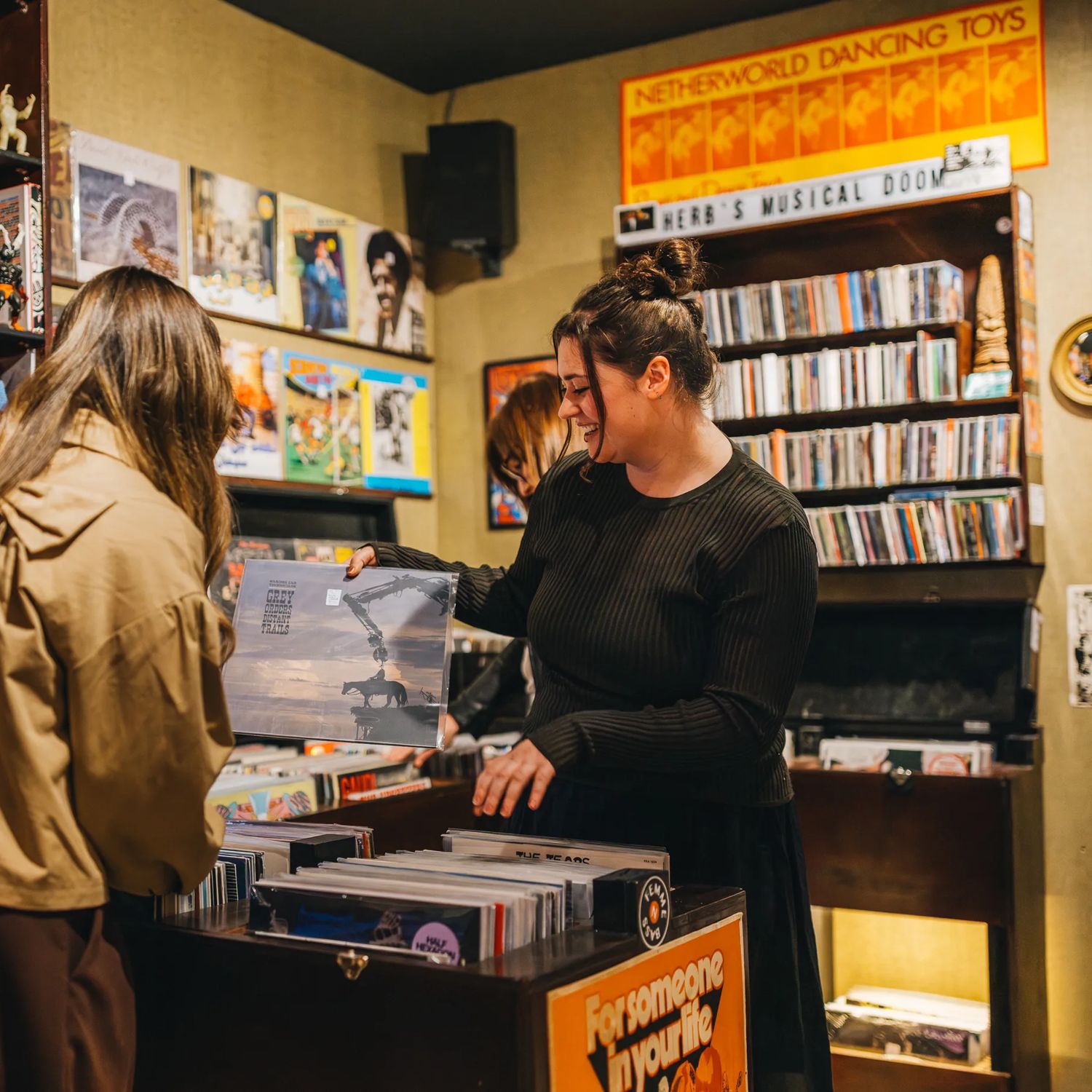 A person stands on one side of a record shop counter while another person behind the counter flips through vinyl records in a bin; shelves and walls around them are filled with records, music posters, and small decorative items under warm indoor lighting.