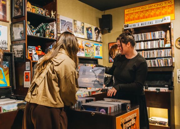 A person stands on one side of a record shop counter while another person behind the counter flips through vinyl records in a bin; shelves and walls around them are filled with records, music posters, and small decorative items under warm indoor lighting.