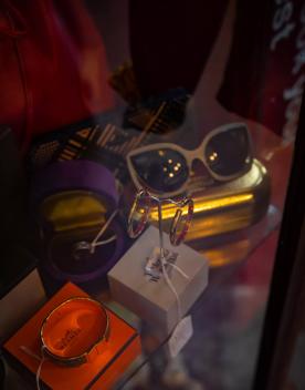 A collection of items on a table — an orange Hermès box with a gold bracelet, a grey Vivienne Westwood box with two hoop earrings on a stand, and a gold glasses case with vintage cream glasses on top.
