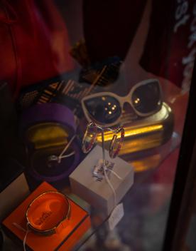 A collection of items on a table — an orange Hermès box with a gold bracelet, a grey Vivienne Westwood box with two hoop earrings on a stand, and a gold glasses case with vintage cream glasses on top.