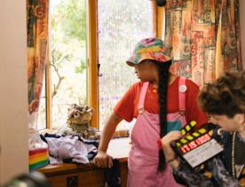 A behind the scenes image from the making of the Wellington-based film, Little Apocolypse with an actor wearing a red tee shirt and pick overalls, leaning on a window sill and another actor wearing a black top holding a clapperboard.