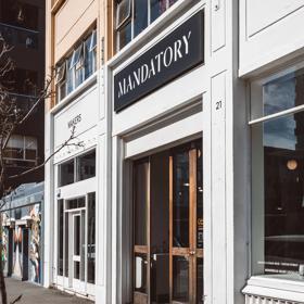 The front facade of Mandatory Menswear Ltd, a men's clothing store in Te Aro, Wellington. It's a white building with wood-framed doors and large windows.