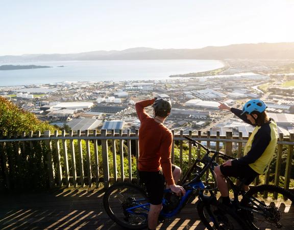 Two cyclists stop at a lookout at Waiu Mountain Bike Park overlooking the city and the harbour below.