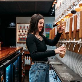 A bartender in Garage Project pours a beer from taps on the wall. Two people are seated at the bar waiting for a drink.