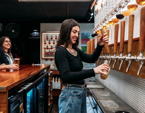 A bartender in Garage Project pours a beer from taps on the wall. Two people are seated at the bar waiting for a drink.
