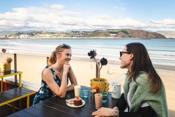 Two people sit and enjoy iced coffees and a dessert on the beach-side patio at Maranui café in Lyall Bay, Wellington.