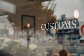 The outside sign of Customs, showing opening times and a glass window into the cafe.