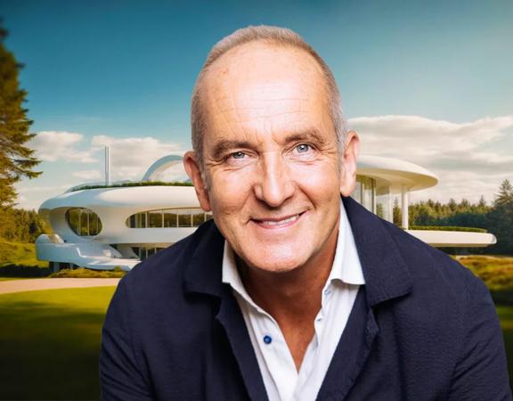 Kevin McCloud, a British designer, writer, and television presenter, smiles in front of a modern three-story white building surrounded by a evergreen forest.
