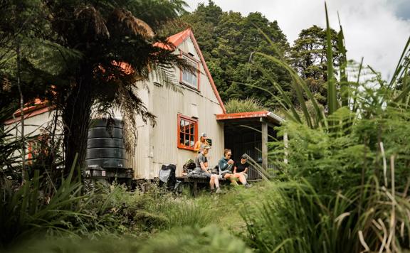 The exterior of Field Hut in Tararua Forest Park. People are sitting on the deck eating. Native trees surround the hut.