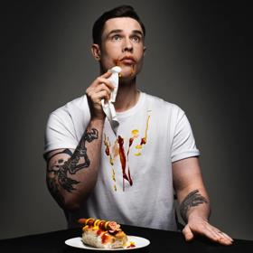 British comedian Ed Gamble dabs his mouth with a napkin, ketchup and mustard on his face and white T-shirt, a hotdog on a plate in front of him.