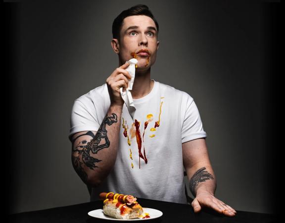 British comedian Ed Gamble dabs his mouth with a napkin, ketchup and mustard on his face and white T-shirt, a hotdog on a plate in front of him.