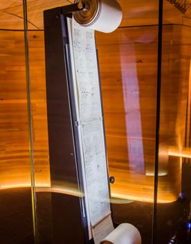 A vertical facsimile is displayed inside a glass case, with a long sheet of densely printed text wound between two rollers at the top and bottom, set against a warm, wood-panelled background.