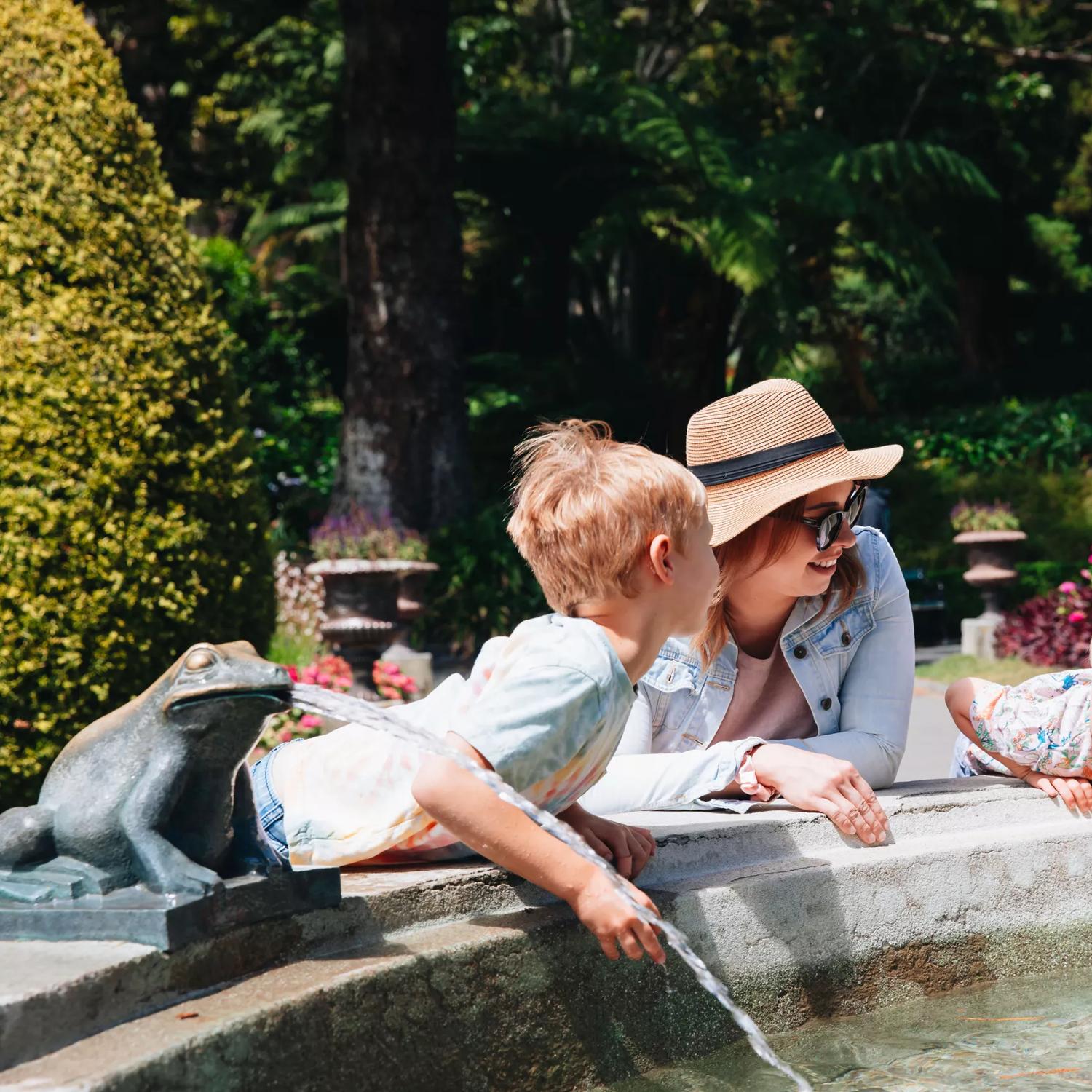 An adult and two kids lean against a public fountain on a sunny day at Wellington Botanic Gardens.