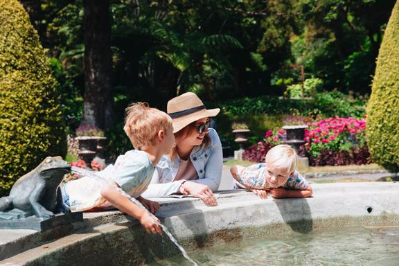 An adult and two kids lean against a public fountain on a sunny day at Wellington Botanic Gardens.